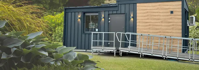 Modernes TinyCareHome-Modulgebäude mit barrierefreier Rampe in parkähnlicher Umgebung. Das kompakte Gebäude kombiniert dunkle Metallfassade mit Holzverkleidung und verfügt über eine Eingangstür mit Beleuchtung.