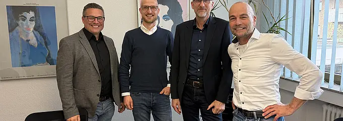 Vier Männer stehen eng nebeneinander in einem hellen Büro- oder Besprechungsraum. Sie lächeln in die Kamera; im Vordergrund ist eine Vertragsmappe auf einem Tisch zu sehen.