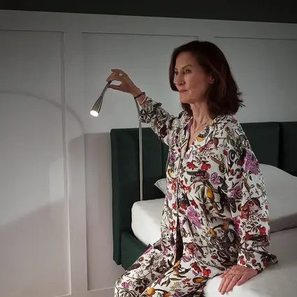 Una mujer está sentada en el borde de la cama, con la lámpara de lectura (accesorio opcional) encendida.