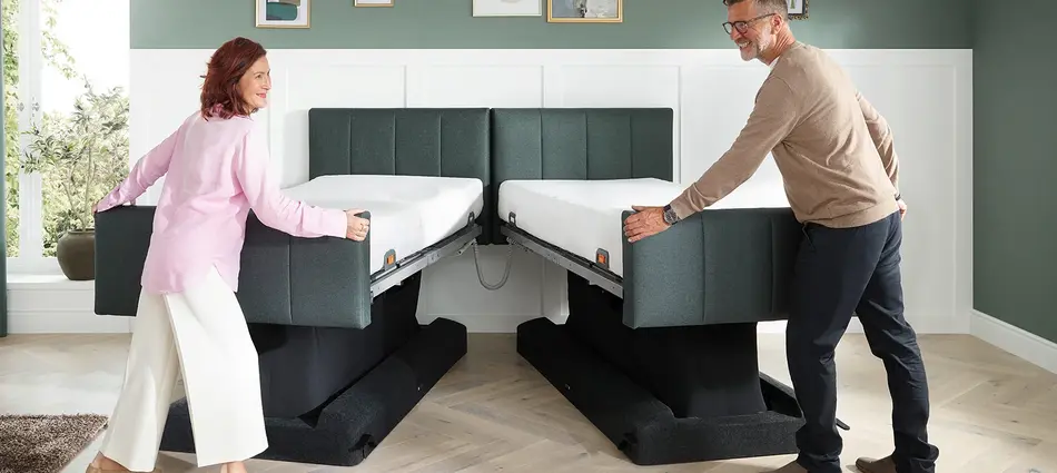 Un hombre y una mujer están de pie a los pies de la cama lifestyle Libra - variante deluxe. Un hombre a la derecha y una mujer a la izquierda están deslizando las 2 camas individuales para hacer una cama doble Libra - variante deluxe 