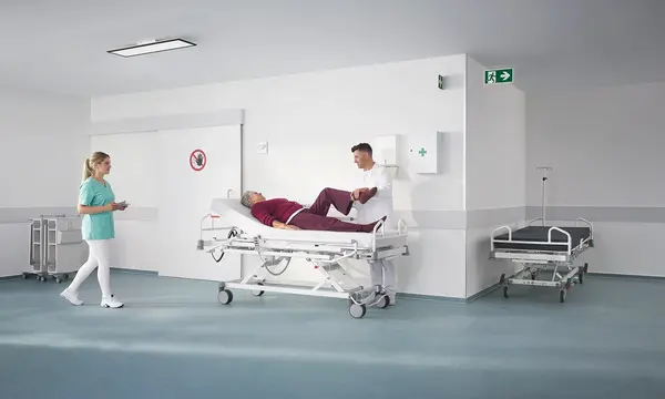 Evario one -sairaalasänky modernissa poliklinikkaympäristössä lääkintälaitteiden kanssa, suunniteltu turvalliseen ja joustavaan potilastyöhön