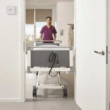 Hoitaja työntää kapeaa leveydeltään 89 cm Evario one -sänkyä oviaukon läpi