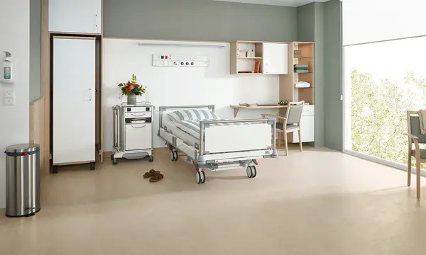Puro hospital bed from Stiegelmeyer in a modern optional service room with bedside cabinet, wardrobe, desk and seating area by the window