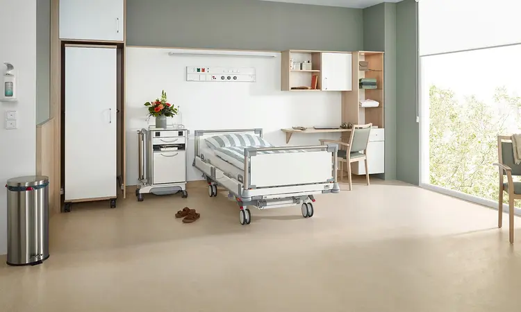 Puro hospital bed from Stiegelmeyer in a modern optional service room with bedside cabinet, wardrobe, desk and seating area by the window