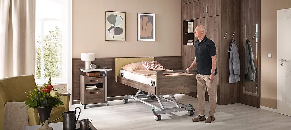 Cama de cuidados Elvido con acabado en madera y ajuste eléctrico de la altura. Con el piecero Primus low y la barandilla, ofrece al paciente seguridad para agarrarse.