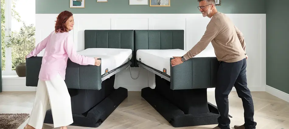 Een man en een vrouw staan aan het voeteneinde van het Libra - variant deluxe lifestylebed. De man rechts en de vrouw links schuiven de twee eenpersoonsbedden in elkaar om er een Libra - variant deluxe tweepersoonsbed van te maken.