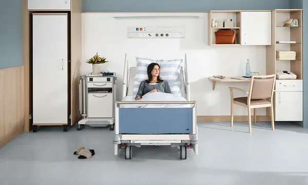 Patientin liegt im Krankenhausbett Evario von Stiegelmeyer in modernem Wahlleistungszimmer mit Schrank, Nachttisch, Schreibtisch und Sitzbereich am Fenster.