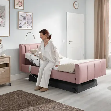 Una mujer está sentada en el borde de la cama y utiliza el mando para ajustar la cama a una altura ideal para levantarse.