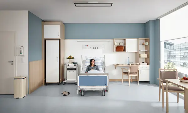 Une patiente est allongée dans un lit hospitalier Evario de Stiegelmeyer dans une chambre moderne pour prestations au choix avec armoire, table de chevet, bureau et coin salon près de la fenêtre.