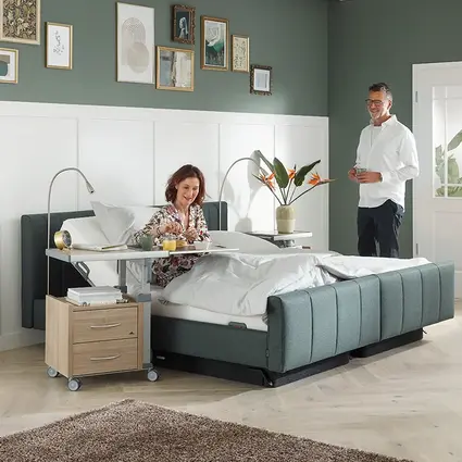 Een vrouw zit in een Libra - variant deluxe lifestyle bed te ontbijten bij een Aparto nachtkastje, terwijl een man naast het bed staat.