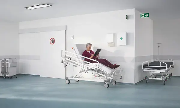 Łóżko szpitalne Evario one w nowoczesnej przychodni ambulatoryjnej z wyposażeniem medycznym, zaprojektowane z myślą o bezpiecznej i elastycznej opiece nad pacjentem.