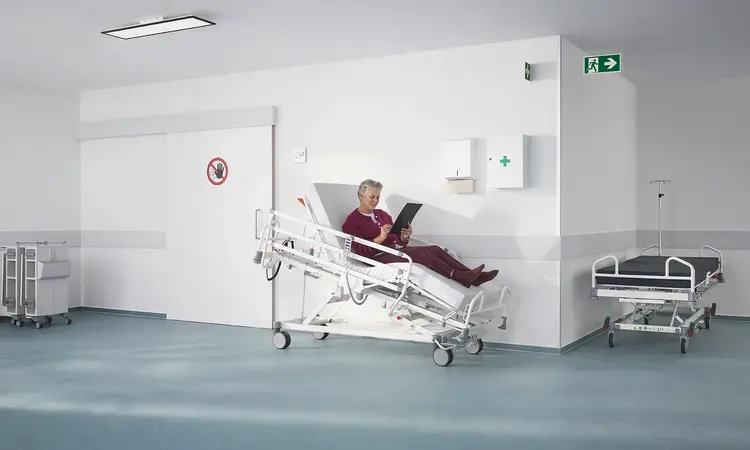 Łóżko szpitalne Evario one w nowoczesnej przychodni ambulatoryjnej z wyposażeniem medycznym, zaprojektowane z myślą o bezpiecznej i elastycznej opiece nad pacjentem.