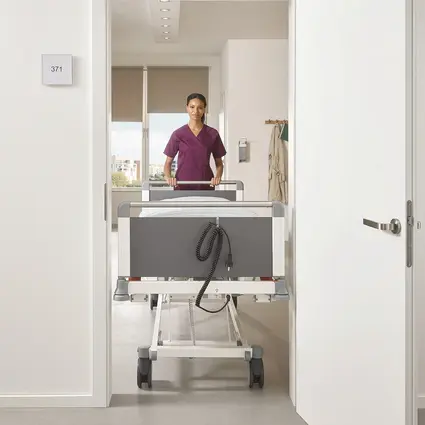 Cuidadora empurra a cama Evario One estreita, com largura de 89cm, através de uma porta