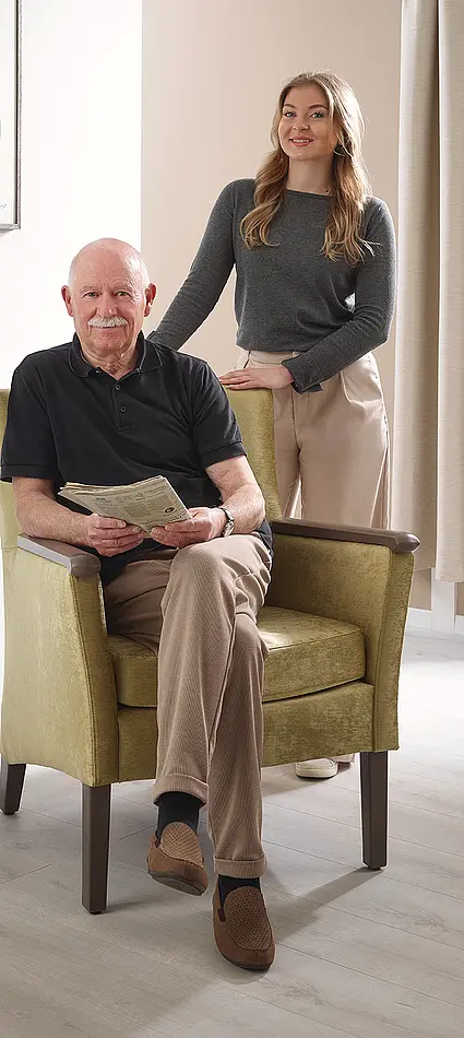 Idoso sentado na cadeira de apoio Luna, acompanhado por uma jovem cuidadora no quarto de cuidados