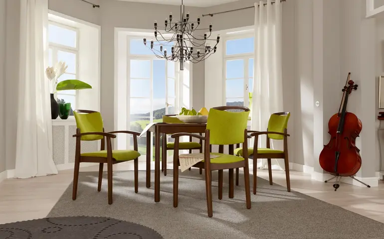 Stapelbarer Stuhl der Serie Fena mit Griffbügel in Holzoptik, stehend an einem Tisch in einem Aufenthaltsraum.