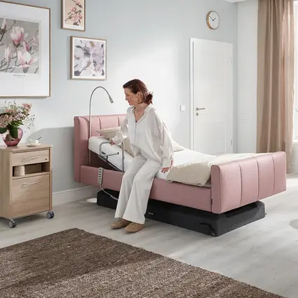 Een vrouw zit op de rand van het bed en gebruikt de handset om het bed op de ideale hoogte te brengen om op te staan.