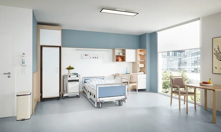 Łóżko szpitalne Evario firmy Stiegelmeyer we współczesnym opcjonalnym pokoju dla pacjenta z szafą, szafką przyłóżkową, biurkiem i miejscem do siedzenia przy oknie.