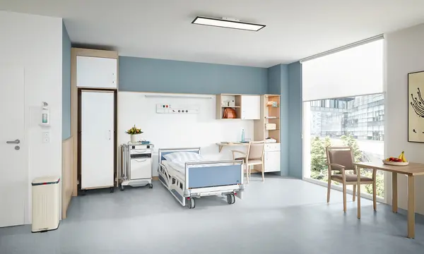 Łóżko szpitalne Evario firmy Stiegelmeyer we współczesnym opcjonalnym pokoju dla pacjenta z szafą, szafką przyłóżkową, biurkiem i miejscem do siedzenia przy oknie.