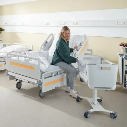 Une patiente est assise sur le bord du lit, le système de protection latérale supérieur relevé l’aide à se lever et à quitter le lit.