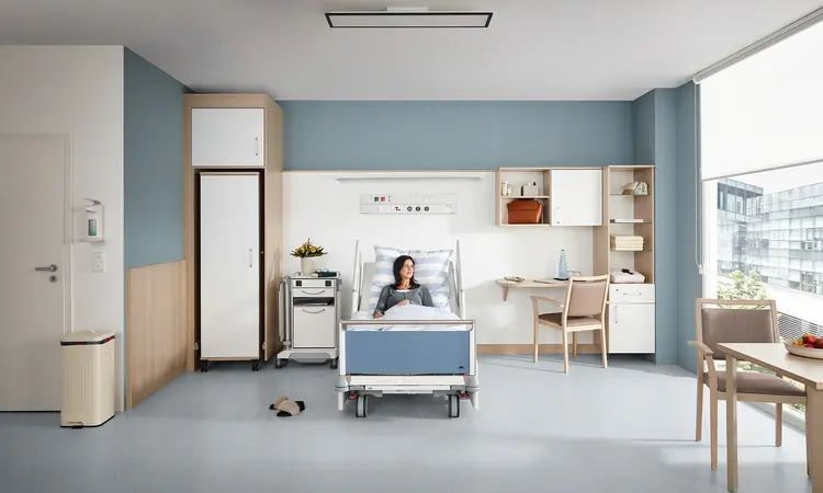 Paciente tumbado en una cama de hospital Stiegelmeyer Evario en una habitación moderna opcional con armario, mesilla de noche, escritorio y zona para sentarse junto a la ventana.