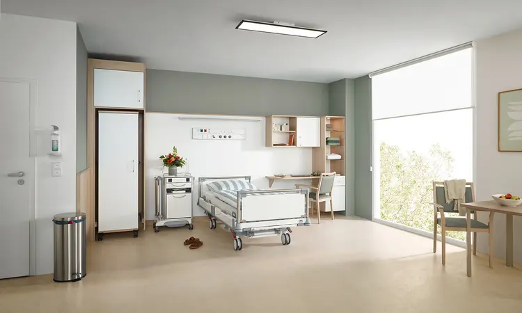 Puro es una cama de hospital de Stiegelmeyer en una moderna habitación de servicios opcionales con mesilla de noche, armario, escritorio y zona para sentarse junto a la ventana.