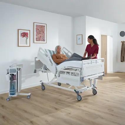 Une patiente est assise dans un lit hospitalier tandis qu’une infirmière la soigne à une hauteur de travail adaptée.